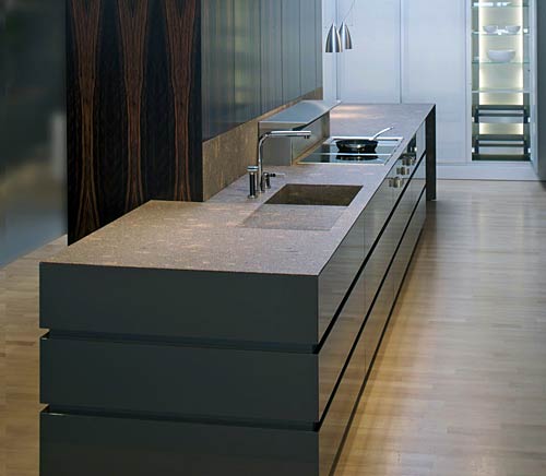 Six metre long island installation as an elegant kitchen architecture of the top class with high glossy lacquered fronts, a natural stone worktop and Makassar veneered tall cabinets; The appliances in this premium kitchen are from Gaggenau.